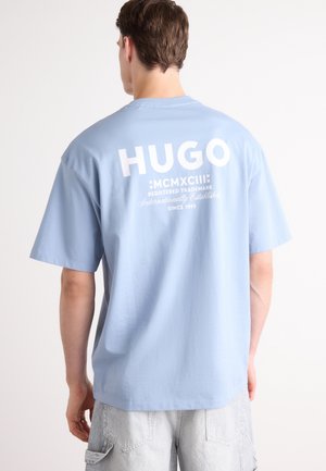 Man wearing light blue T-shirt with white "HUGO" logo and text on back, paired with light gray striped shorts.