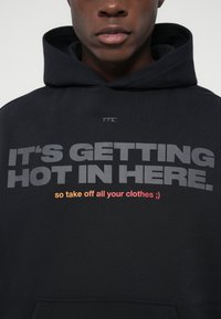 Άνδρας με μαύρο φούτερ που έχει εκτυπωμένο μπροστά το κείμενο "ΓΙΝΕΤΑΙ ΖΕΣΤΟ ΕΔΩ. οπότε βγάλε όλα σου τα ρούχα ;)".