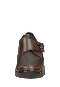 Zapato marrón de cuero tipo mocasín con una correa de velcro, forma redondeada y suela de goma negra. Presenta un acabado suave y texturizado.