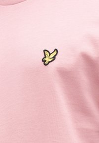 Vaaleanpunainen puuvillainen T-paita, jossa vasemmalla rintakehän alueella pieni mustakultainen lintukirjailu, sileä tekstuuri ja pyöreä pääntie.