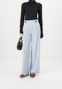 Světle modré kalhoty s širokými nohavicemi a předními záhyby, černé knoflíky, v kombinaci s černým topem s dlouhým rukávem a balerínami s leopardím potiskem, držící černou kabelku.