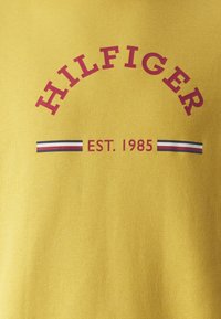 Žlté bavlnené tričko s červeným zakriveným logom "HILFIGER" a textom "EST. 1985", s pruhovaným akcentom v námorníckej modrej, červenej a bielej pod ním.