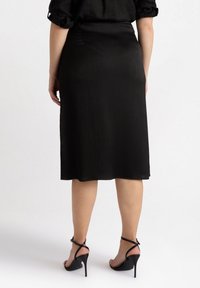 Zwarte geplisseerde midi rok, gladde textuur, licht taps toelopend silhouet, met een hoge taille en een zoom die net onder de knie eindigt. Zwarte hakken sandalen.