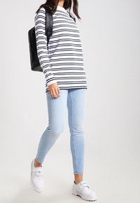 Femme marchant vêtue d'un haut à manches longues blanc et noir à rayures, jean skinny bleu clair, chaussures blanches et un sac à dos noir.