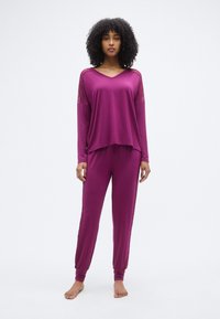 Σετ με μακρυμάνικη μπλούζα με V λαιμόκοψη και παντελόνι σε ζωντανό μωβ χρώμα, με δαντελένιες λεπτομέρειες στα μανίκια και στενές μανσέτες στον αστράγαλο για άνετη εφαρμογή.