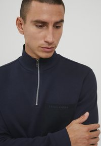 Tricou cu fermoar la gât de culoare marină, cu un guler înalt, dotat cu un buzunar texturat și detalii brodate "CASUAL FRIDAY". Textură a materialului neted.
