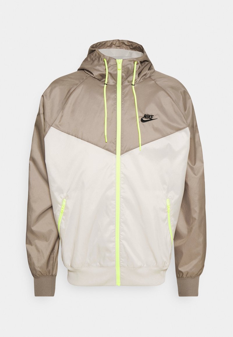 Veste à capuche Nike avec des panneaux beige et crème, en matériau léger, fermeture éclair jaune fluo et logo sur la poitrine gauche. Poignets et ourlet élastiques.