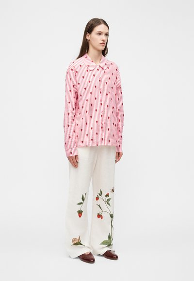 Woman wearing a pink and white striped button-up shirt with small strawberries, white pants with strawberry and snail prints, and brown shoes.