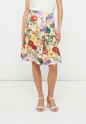 Femme portant une jupe longueur genou à imprimé floral coloré, associée à des sandales blanches à talons et un top blanc sans manches.