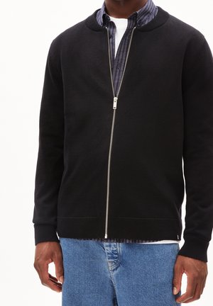 Mężczyzna ubrany w czarny kardigan na zamek, na częściowo rozpiętą, w paski koszulę zapinaną na guziki oraz niebieskie dżinsy, stojący na jednolitym tle.