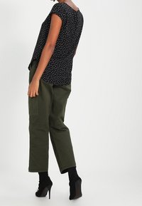 Czarna bluzka w groszki z krótkimi rękawami, z tylnym zapięciem na guziki; w zestawie z oliwkowozielonymi szerokimi spodniami; czarne szpilki.