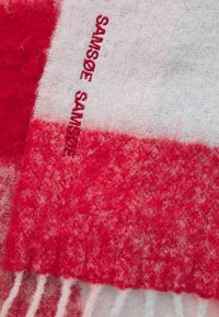 Close-up van rood en wit zacht textiel met "SAMSOE SAMSOE" geborduurde in rode draad op een wit gedeelte.