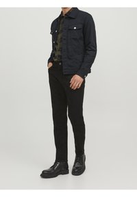 Černá džínová bunda s dvěma náprsními kapsami přes zelenou a černou kostkovanou košili, doplněná černými slim-fit kalhotami a černými botami.