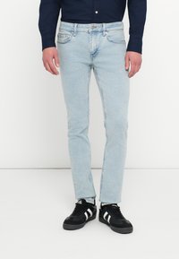 Mies, joka käyttää vaaleansinisiä slim fit -farkkuja, tummaa pitkähihaista paitaa ja mustia valkoraidallisia lenkkareita, seisoo yksinkertaisella taustalla.