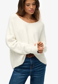 Witte oversized trui met een brede halslijn, geribbelde manchetten en een zachte textuur, gecombineerd met donkerblauwe spijkerbroeken en een gevlochten bruine riem.