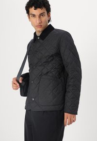 Casaco acolchoado preto com colarinho em algodão, apresentando botões de pressão e um bolso no peito. Modelo segurando uma bolsa mensageiro escura, posicionado contra um fundo branco.