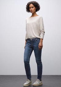 Vaaleanbeige pitkähihaisessa neuleessa, joka on yhdistetty sinisiin skinny-farkkuihin. Farkuissa on huomaamaton etutaskujen malli ja harmaat lenkkarit.