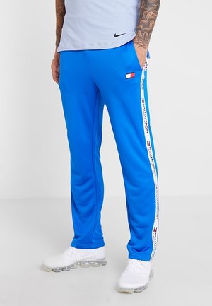 Mężczyzna w jasnoniebieskich spodniach dresowych Tommy Sport z białymi paskami po bokach i logo, białych trampkach oraz jasnoszarej koszulce Nike.