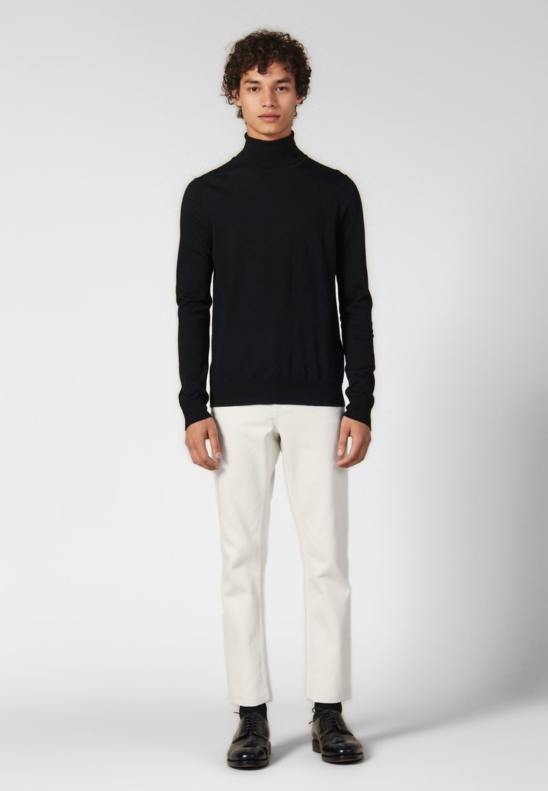 Maglione nero a collo alto in una vestibilità slim, abbinato a pantaloni color crema e scarpe nere. Il tessuto sembra liscio, con maniche attillate.