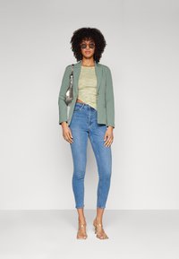 Blazer verde claro sobre una blusa de patrón dorado, combinado con jeans ajustados azules y sandalias de tacón transparente. La modelo sostiene un bolso plateado.