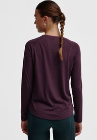 Langärmliges, tiefpurpurnes Sportoberteil mit abgerundetem Saum und Raglanärmeln, aus strukturiertem Stoff. Getragen mit dunkelgrünen Leggings.