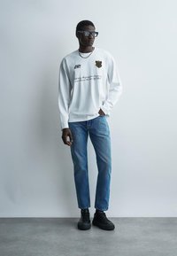 Vīrietis, kas valkā melnas brilles, baltu garām piedurknēm kreklu ar tekstu un ģerboni, zilas džinsas un melnas sporta kurpes, stāv pie vienkāršas gaiši pelēkas sienas.