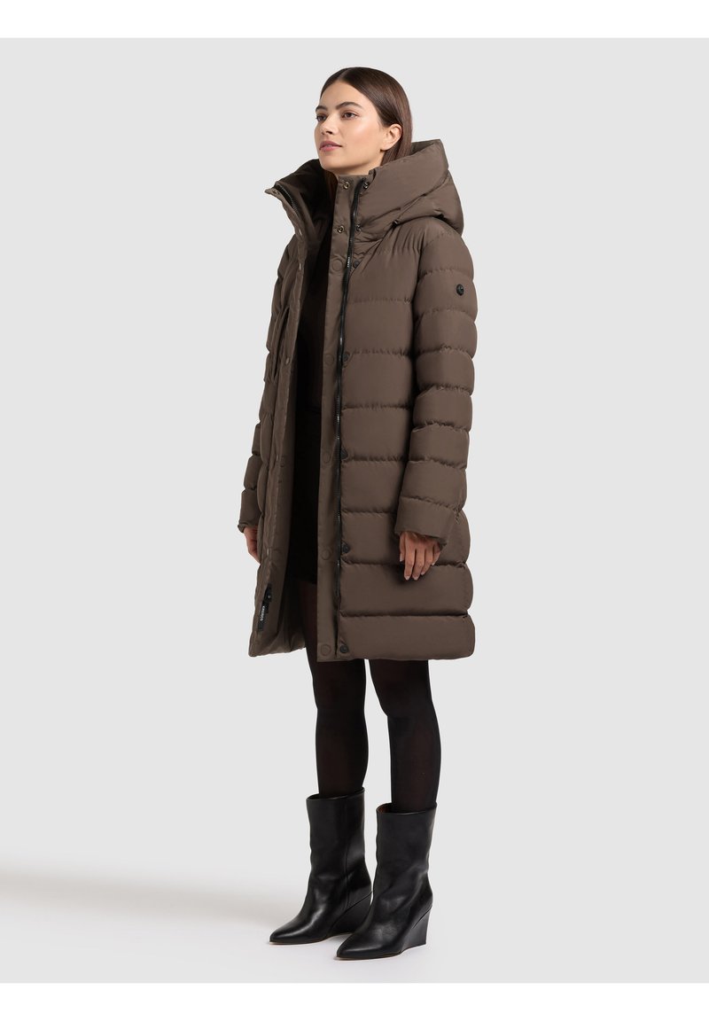 Hnědý prošívaný kabát s vysokým límcem, vybavený knoflíkovou sponou vpředu a žebrovanou texturou. Nositelný s černými kozačkami ke kolenům a tmavým oblečením.