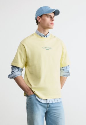 Tânăr îmbrăcat într-un tricou Calvin Klein galben deschis peste o cămașă dungată cu nasturi, blugi albaștri deschiși și o șapcă albastră asortată, cu mâinile în buzunare.