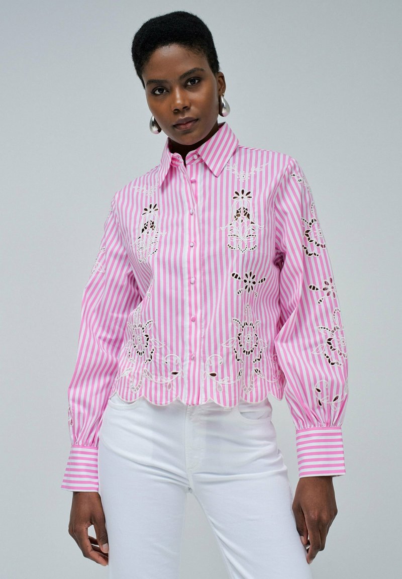 Blouse rayée rose et blanche avec broderies florales et fermeture à boutons. Elle présente des manches bouffantes et un ourlet en forme de vagues.