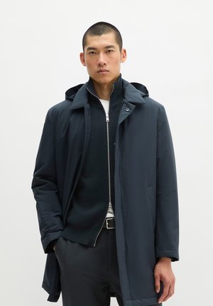 Mann trägt einen dunkelblauen Kapuzenmantel über einer schwarzen Jacke mit Reißverschluss und einem weißen Hemd, stehend vor einem einfarbigen weißen Hintergrund.