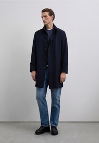 Mand iført en marineblå frakke over en blå sweater og lyseblå jeans, der står foran en ensfarvet lys grå væg og betongulv.
