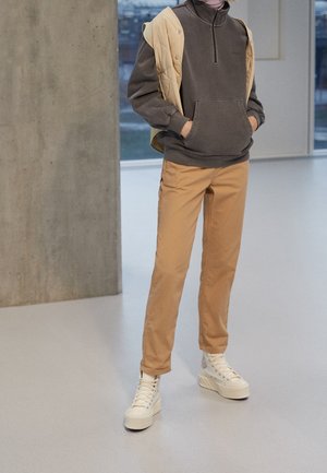 Person trägt braunes Sweatshirt mit Viertel-Reißverschluss, hellbraune Hose, cremefarben gesteppte Weste über den Schultern und weiße Plateau-High-Top-Sneaker in Innenräumen.