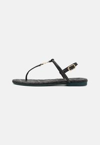 Sorte flate sandaler med vevet tekstur, tanga-stil stropp, justerbar ankelspenne og en mørkegrønn såle. Gulldetaljer.