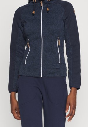 Granatowa kurtka zapinana na zamek z kapturem, białym zamkiem i zamkami w kieszeniach, noszona z dopasowanymi granatowymi spodniami.