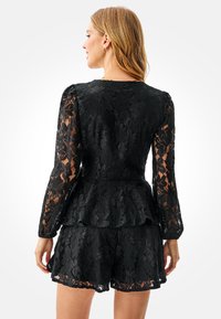 Schwarzes Spitzen-Peplum-Top mit langen Ärmeln, das eine florale Struktur und durchsichtige Akzente aufweist. Das Design beinhaltet einen mehrlagigen Saum und eine taillierte Silhouette.