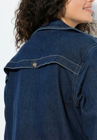 Modrá džínová bunda s špičatým límcem, velkou zadní vsadkou a kovovým zatrhávacím zapínáním. Látka má hladkou strukturu s jemnými šicími detaily.