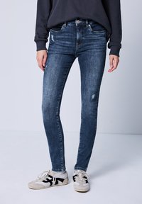 Kapeet siniset skinny farkut, joissa on kulunut laikku. Yhdistetty vaaleanharmaisiin ja mustiin tennareihin, neutraalin taustan edessä.