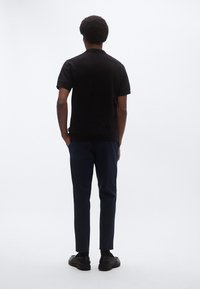 Μαύρο πόλο πουκάμισο με κοντά μανίκια, μπλε παντελόνι, φιγούρα που στέκεται από πίσω σε ουδέτερο φόντο, με απλό σχέδιο και υφασμάτινη υφή.