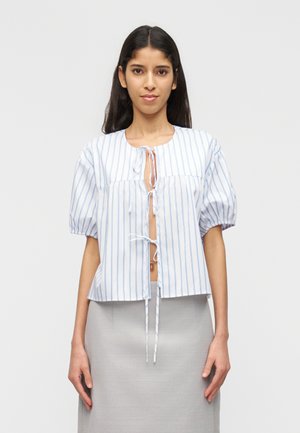 Kobieta ubrana w biały krótki top w niebieskie pionowe paski, z bufiastymi rękawami i wiązaniem z przodu, w połączeniu z wysokim szarym spódnicą.