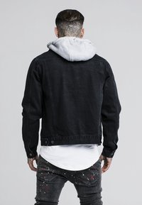Schwarze Jeansjacke mit einem grauen Kapuzenpullover darunter. Besitzt einen klassischen Schnitt, Vordertaschen und dezente Nähte. Getragen über einem weißen Hemd.