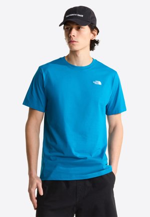 Giovane uomo che indossa una maglietta blu The North Face e un cappello nero, in piedi con una mano in tasca, che guarda leggermente di lato.