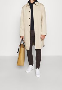 Beige kappa med krage och bruna knappar, bärs över en marinblå hoodie. Bruna byxor och vita sneakers. Håller en beige väska med ett grått handtag.
