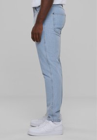 Jeans in denim blu chiaro con una vestibilità slim, caratterizzati da un design a cinque tasche e cuciture sottili. Indossati con sneaker bianche.
