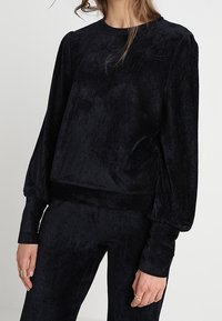 Svart plyschpullover med långa, vida ärmar, rund halsringning och en mjuk, texturerad yta. Tight vid midjan för en kortad silhuett.