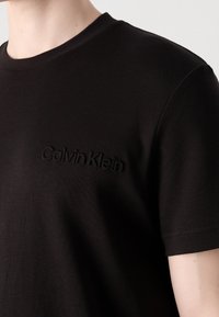 Gros plan d'une personne portant un t-shirt noir Calvin Klein avec le nom de la marque embossé sur le côté gauche de la poitrine.