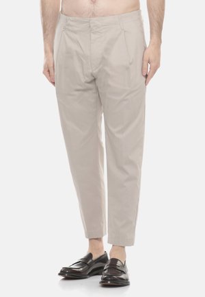 Pantaloni da uomo beige su misura con pieghe frontali, indossati con mocassini in pelle marrone scuro a piedi nudi.