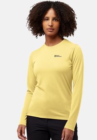Junge Frau mit lockigem Haar, die ein langärmliges gelbes Jack Wolfskin-Shirt und schwarze Hosen trägt, steht vor einem einfarbigen Hintergrund.