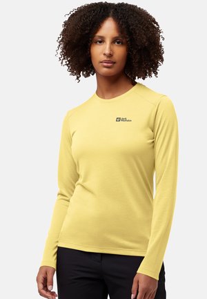 Giovane donna con capelli ricci che indossa una camicia gialla a maniche lunghe della Jack Wolfskin e pantaloni neri, in piedi di fronte a uno sfondo semplice.