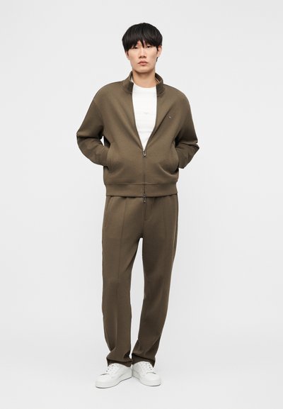 Νεαρός άνδρας που φοράει χακί μπουφάν με φερμουάρ και παντελόνι σετ, μαζί με λευκά αθλητικά, στέκεται με τα χέρια στις τσέπες μπροστά σε λευκό φόντο.