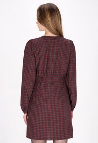 Robe à carreaux rouge et vert foncé avec des manches longues, taille cintrée et poignets froncés. Tissu lisse et silhouette droite.